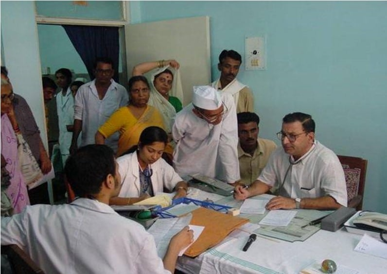 Dr. Ulhas Jajoo conducting an outpatient clinic at Kasturba Hospital, MGIMS Sevagram, with residents and patients gathered around the desk.