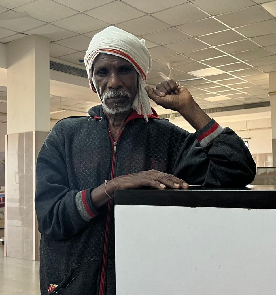 A caregiver husband stands at the nursing station in the palliative care ward at MGIMS Sevagram, dressed in traditional rural clothing
