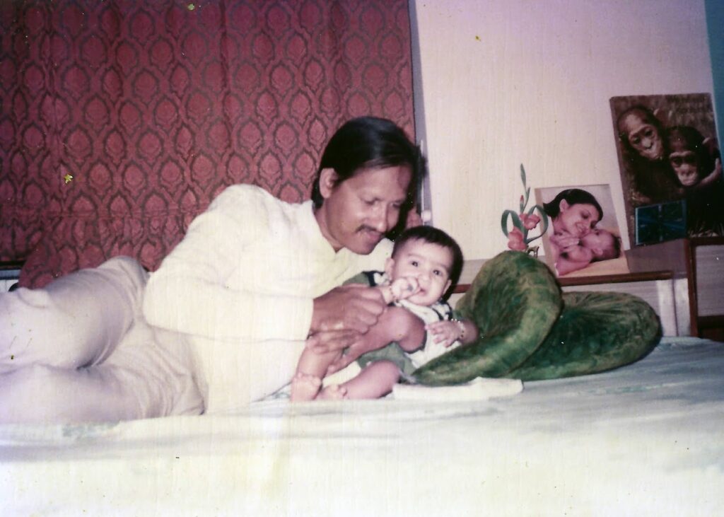 A vintage 1986 photograph of a young father lying on a bed and smiling down at his infant son, Ashwini, who is looking toward the camera