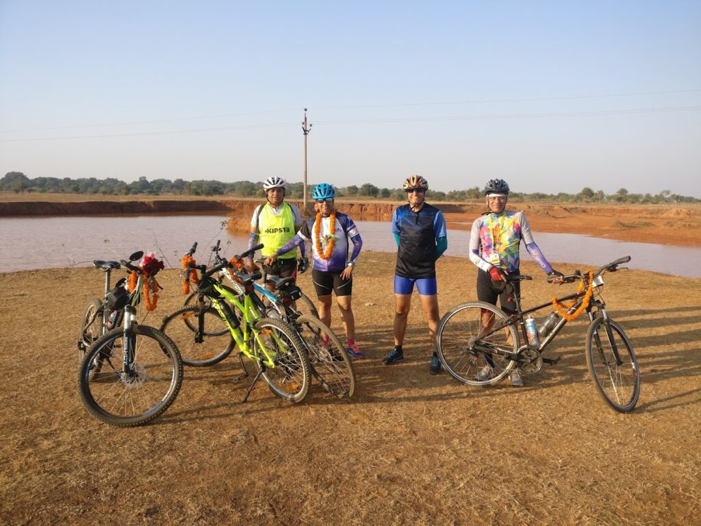 Dr. S.P. Kalantri with cycling companions beside a lake on the Raipur–Bastar route, Chhattisgarh, 2018. Marigold garlands on the bicycles — a welcome from local hosts.