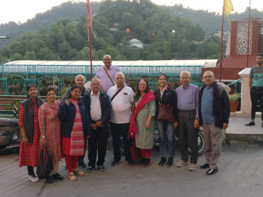 Ten people, five men and five women, standing together on a terrace overlooking a wooded hillside at dusk, with a hotel building and coloured railing behind them