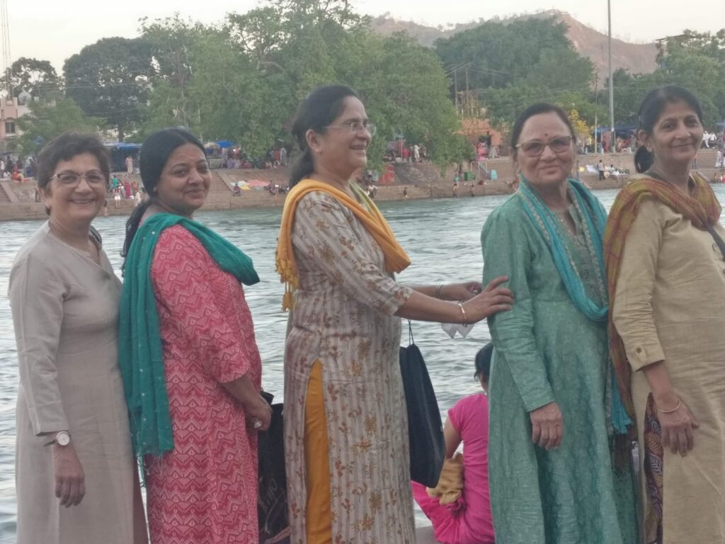 Five women in traditional Indian dress standing together on the banks of the Ganges river at Rishikesh, with trees and a ghat in the background