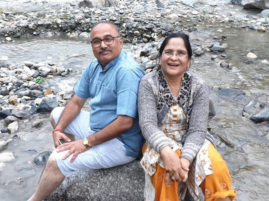 A man and woman sitting back to back on a large rock in a rocky riverbed, smiling, in the Himalayas